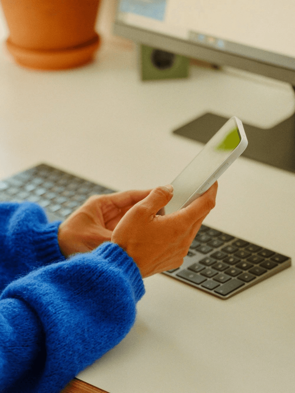 A person in a blue sweater holding a phone at their desk.